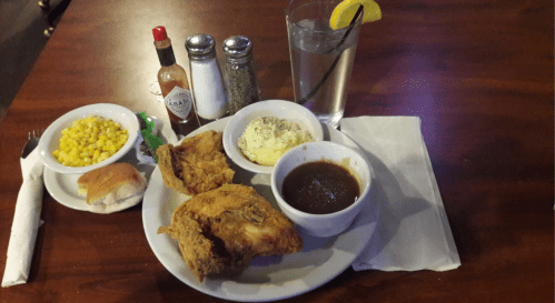 A plate of fried chicken with sides of corn, mashed potatoes, gravy, a roll, and a glass of water with lemon.