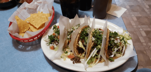 A plate of three tacos topped with lettuce and tomatoes, alongside a basket of tortilla chips.