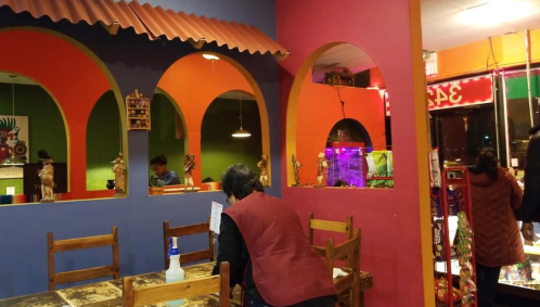 Colorful restaurant interior with arched walls, wooden tables, and decorative figures. A person is seen at the counter.