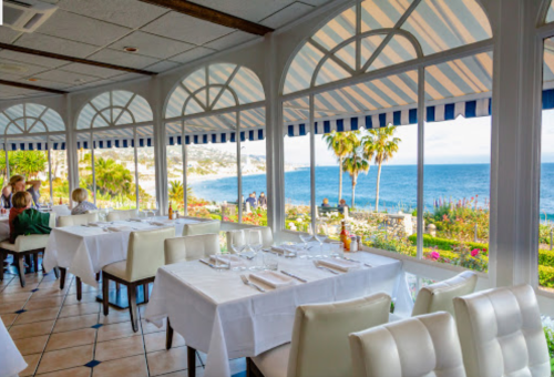 Bright restaurant interior with white tables, overlooking a scenic ocean view through large windows.