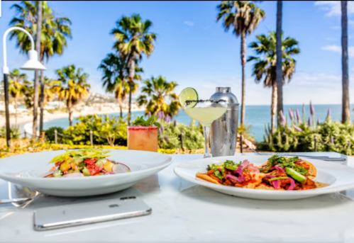 A vibrant outdoor dining scene with two colorful plates of food, cocktails, and palm trees by the beach in the background.