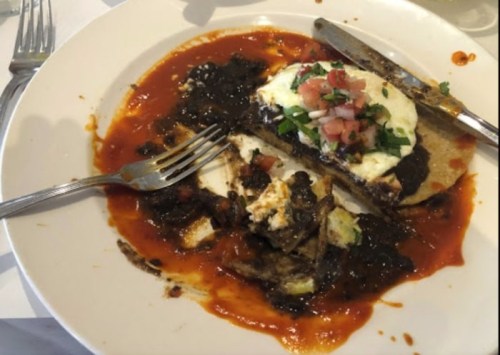 A plate of food featuring a tortilla topped with eggs, black beans, and salsa, surrounded by a red sauce. Fork and knife included.