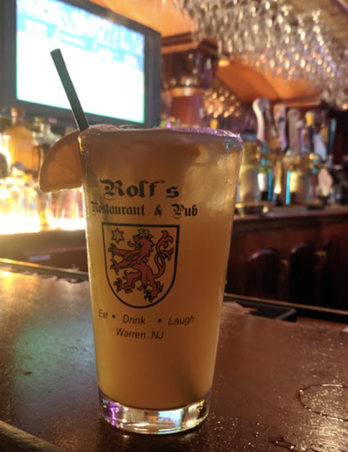 A glass of orange drink with a slice of orange, sitting on a bar counter with a blurred background of bottles.