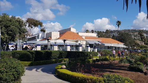 A restaurant with a red-tiled roof surrounded by greenery and palm trees, under a blue sky with fluffy clouds.