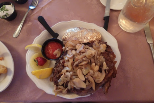 A plate of food featuring a large steak topped with mushrooms and onions, served with a baked potato and lemon wedges.