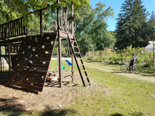 A wooden playground structure beside a path, with a person riding a bike in a sunny, green park setting.