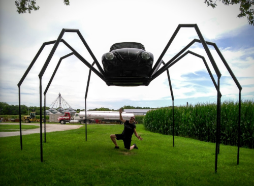 A person kneels in awe beneath a giant spider sculpture made from a car and metal legs, set against a rural backdrop.