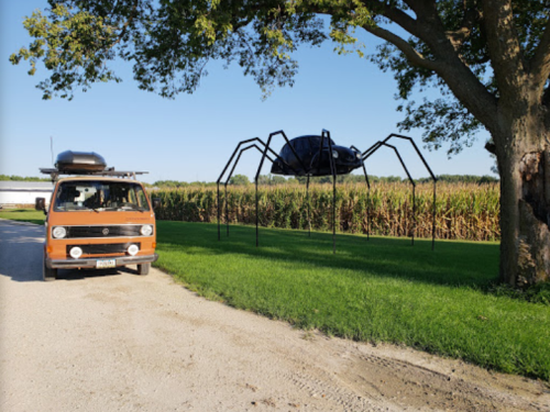 A vintage orange van parked near a giant spider sculpture made from a car, set against a cornfield backdrop.