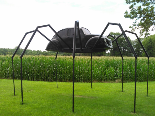 A large sculpture of a spider with a car body on top, set in a grassy area near a cornfield.