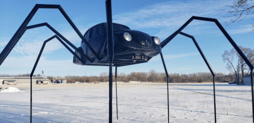 A large sculpture of a black car with spider legs, elevated above a snowy field under a clear blue sky.