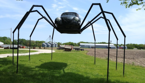 A large sculpture of a spider made from a car, elevated on metal legs, set in a grassy field with farm buildings in the background.