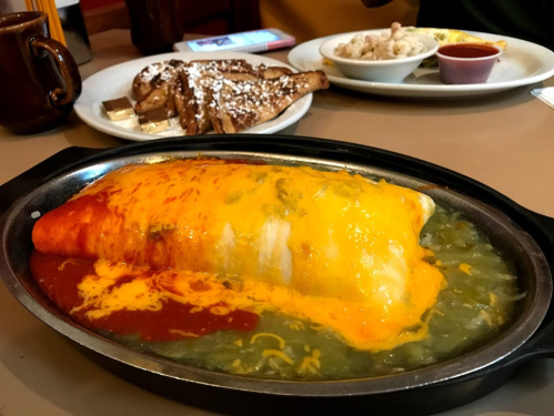A large, cheesy burrito topped with red sauce and green sauce, served on a sizzling plate with sides in the background.