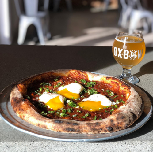 A close-up of a pizza topped with eggs and green onions, alongside a glass of beer on a table.