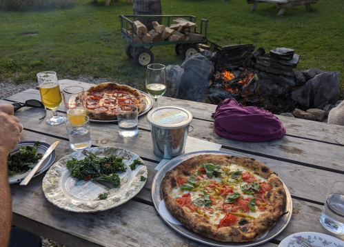 A rustic outdoor table with two pizzas, a salad, drinks, and a fire pit in the background.