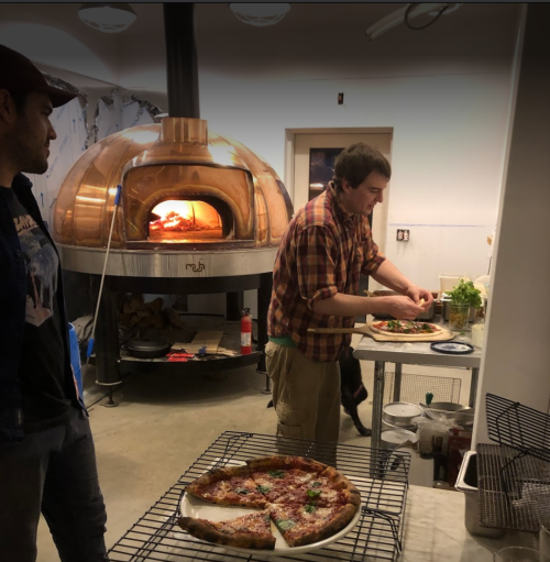 A man prepares pizza in front of a wood-fired oven while another man watches in a cozy kitchen setting.