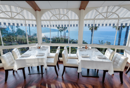 Bright restaurant interior with white tables and chairs, overlooking a scenic ocean view through large windows.