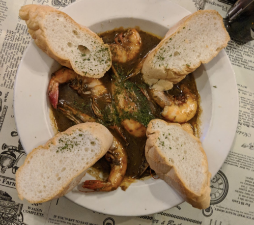 A plate of shrimp in a dark sauce, garnished with parsley, surrounded by slices of bread on a patterned tablecloth.