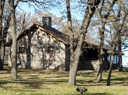 A rustic stone cabin surrounded by trees in a park, with a clear blue sky above and picnic tables in the background.