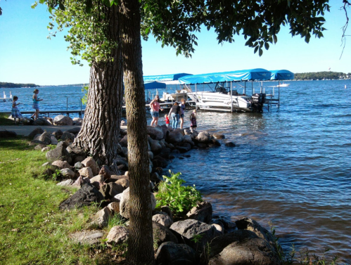 A sunny lakeside scene with people by the water, a boat docked, and trees lining the shore.