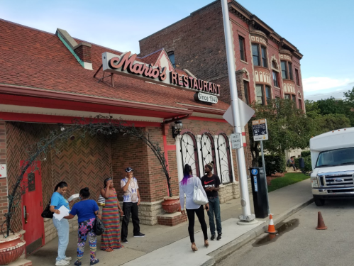 A group of people stands outside Mario's Restaurant, a brick building with a sign, on a sunny day.