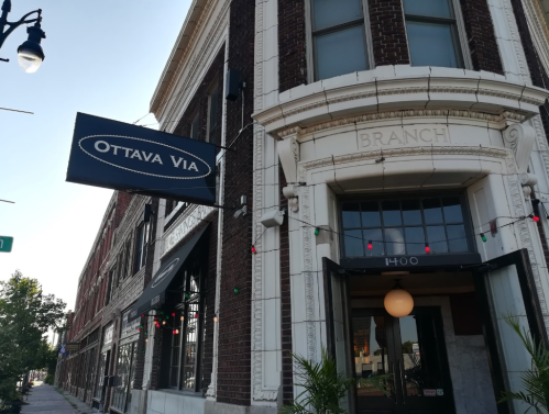 Exterior of a brick building with a sign reading "Ottava Via" and decorative lights, showcasing a welcoming entrance.