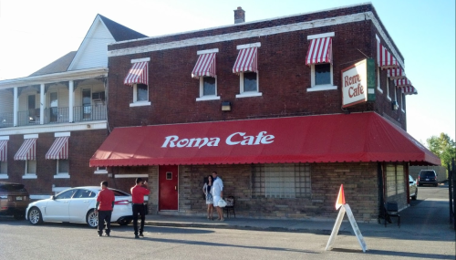 Exterior of Roma Cafe, a brick building with red and white striped awnings, featuring two people outside and a sign.