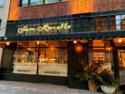 Exterior of San Morello restaurant, featuring a neon sign and warm lighting, with decorative plants at the entrance.