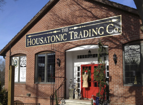 A brick building with a sign reading "The Housatonic Trading Co." and a red door, featuring a mannequin in the window.