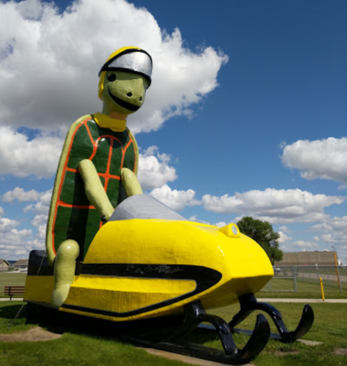 A large turtle statue wearing a helmet, riding a yellow snowmobile against a blue sky with fluffy clouds.