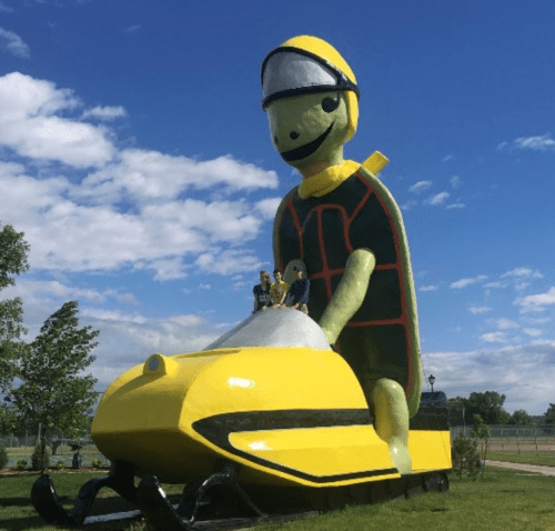 A large green statue of a person on a yellow snowmobile, with three people sitting on it, against a blue sky.