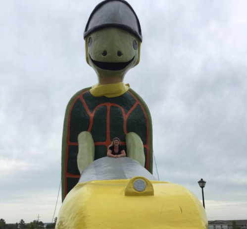 A large turtle statue with a smiling face, featuring a person sitting on its back, under a cloudy sky.