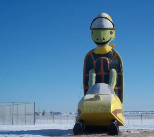 A large snowmobile statue with a cartoonish driver, set against a clear blue sky and snowy landscape.