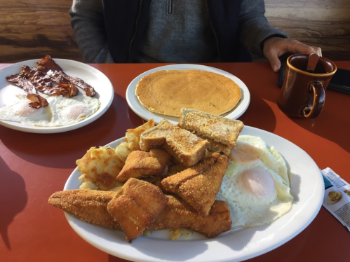 A hearty breakfast plate with fried fish, eggs, toast, and hash browns, alongside a plate of bacon and pancakes.