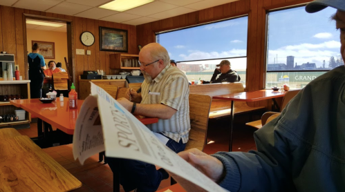 A diner scene with wooden decor, featuring patrons at tables and a man reading a newspaper in the foreground.