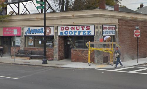 A street view of a donut and coffee shop with construction scaffolding outside, near a bus stop and other storefronts.