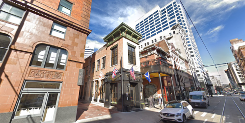 Historic buildings line a city street, featuring flags and a mix of architectural styles under a bright blue sky.