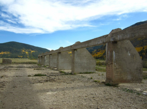 Ruins of a concrete structure in a barren landscape, surrounded by mountains and autumn foliage under a blue sky.