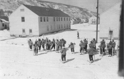 A group of skiers in winter gear gathers on a snowy street near white buildings in a mountainous area.