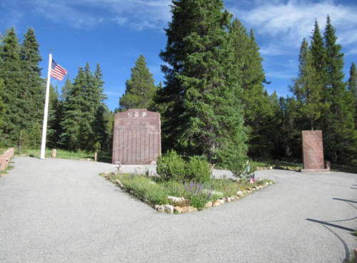 A gravel path leads through a forest, featuring two stone monuments and an American flag in the background.