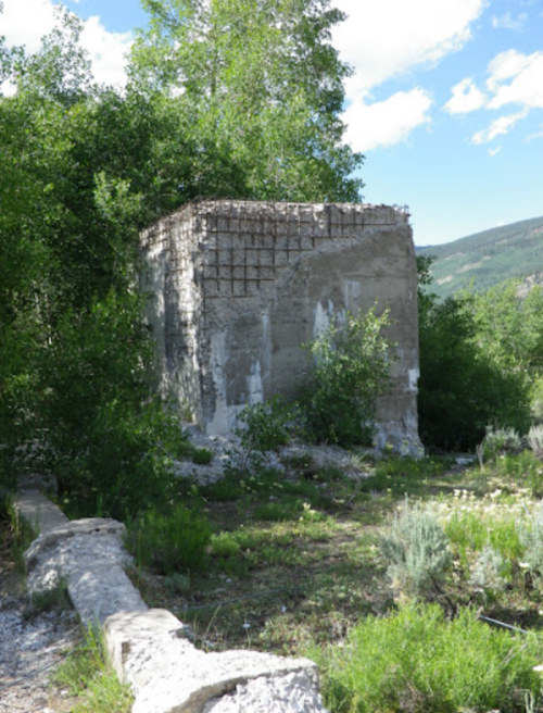 Abandoned concrete structure surrounded by greenery and trees, with a mountainous landscape in the background.