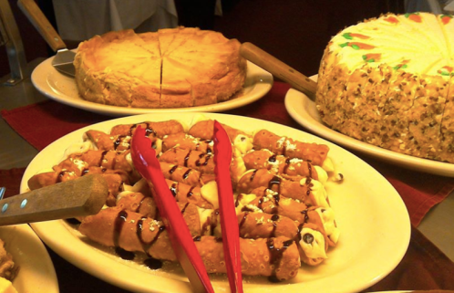 A platter of cannoli drizzled with chocolate sauce, with cheesecake and a nut cake in the background.