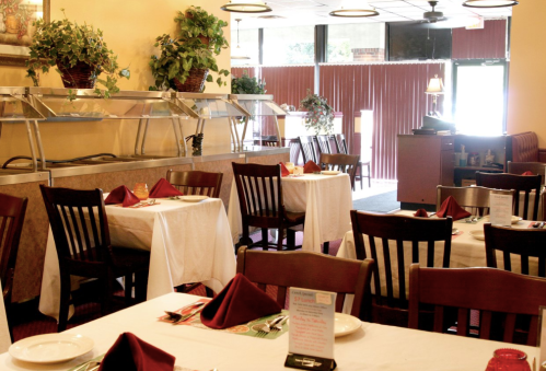 A cozy restaurant interior with white tablecloths, wooden chairs, and a buffet area, featuring plants and warm lighting.