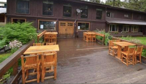 A wooden deck with tables in front of a rustic building surrounded by greenery.
