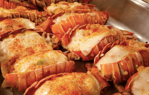 A close-up of baked lobster tails topped with melted cheese and sprinkled with seasoning.
