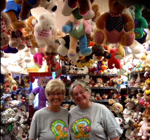 Two smiling women stand in a colorful room filled with hanging stuffed animals and teddy bears.