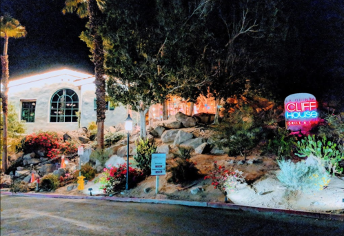 A lit restaurant exterior at night, surrounded by rocks and colorful plants, with a sign reading "Cliff House."