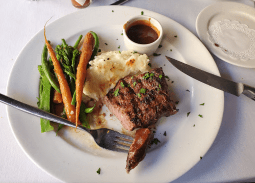 A plate with grilled steak, mashed potatoes, green beans, and carrots, accompanied by a small bowl of sauce.