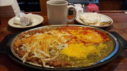 A sizzling plate of beans, hash browns, cheese, and eggs, served with tortillas and a cup of coffee.