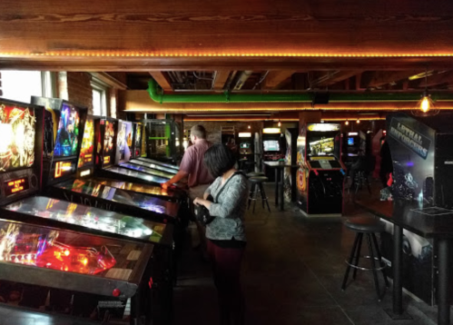 A retro arcade with pinball machines and players enjoying the games in a dimly lit space.