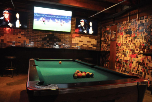 A dimly lit pool hall with a green table, colorful balls, and two screens showing a sports game on the wall.